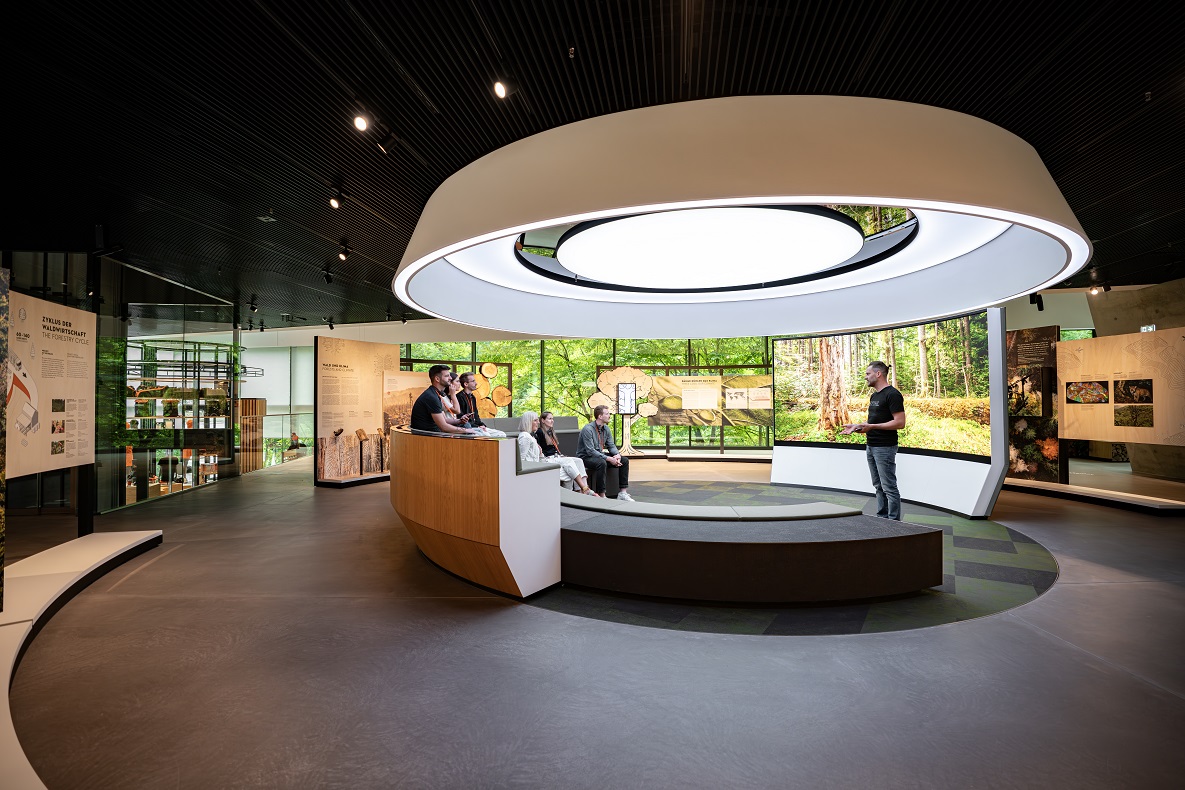 Moderner Präsentationsraum in der STIHL Markenwelt mit Besuchern, großem Lichtkreis und Naturmotiven im Hintergrund.